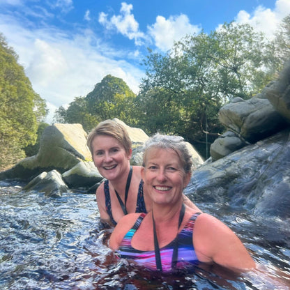 Two women in swimsuits sitting in the river surrounded by rocks and trees.