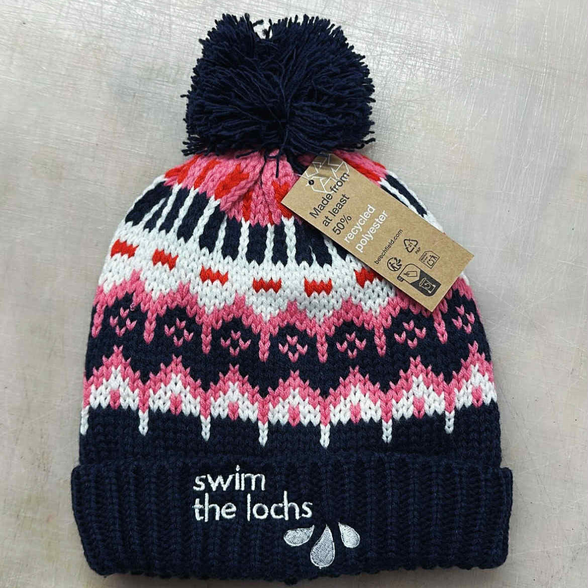 Knitted beanie with a pom-pom and 'swim the lochs' branding on a light background